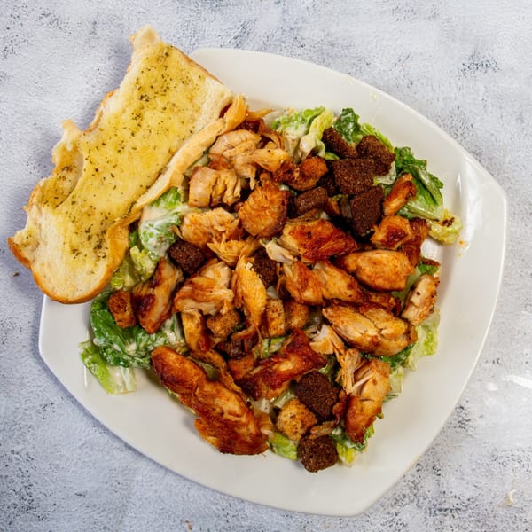 Chicken Caesar salad with croutons, dressing, and a side of garlic bread on a white plate.