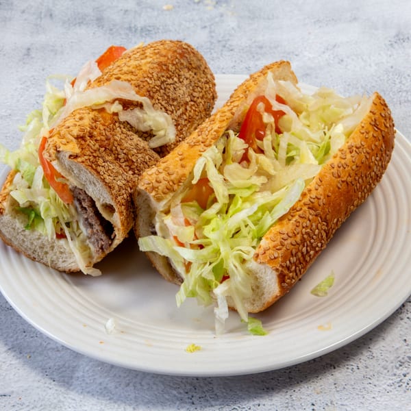 Two sesame seed hoagies filled with meat, lettuce, tomatoes, and onions on a white plate.