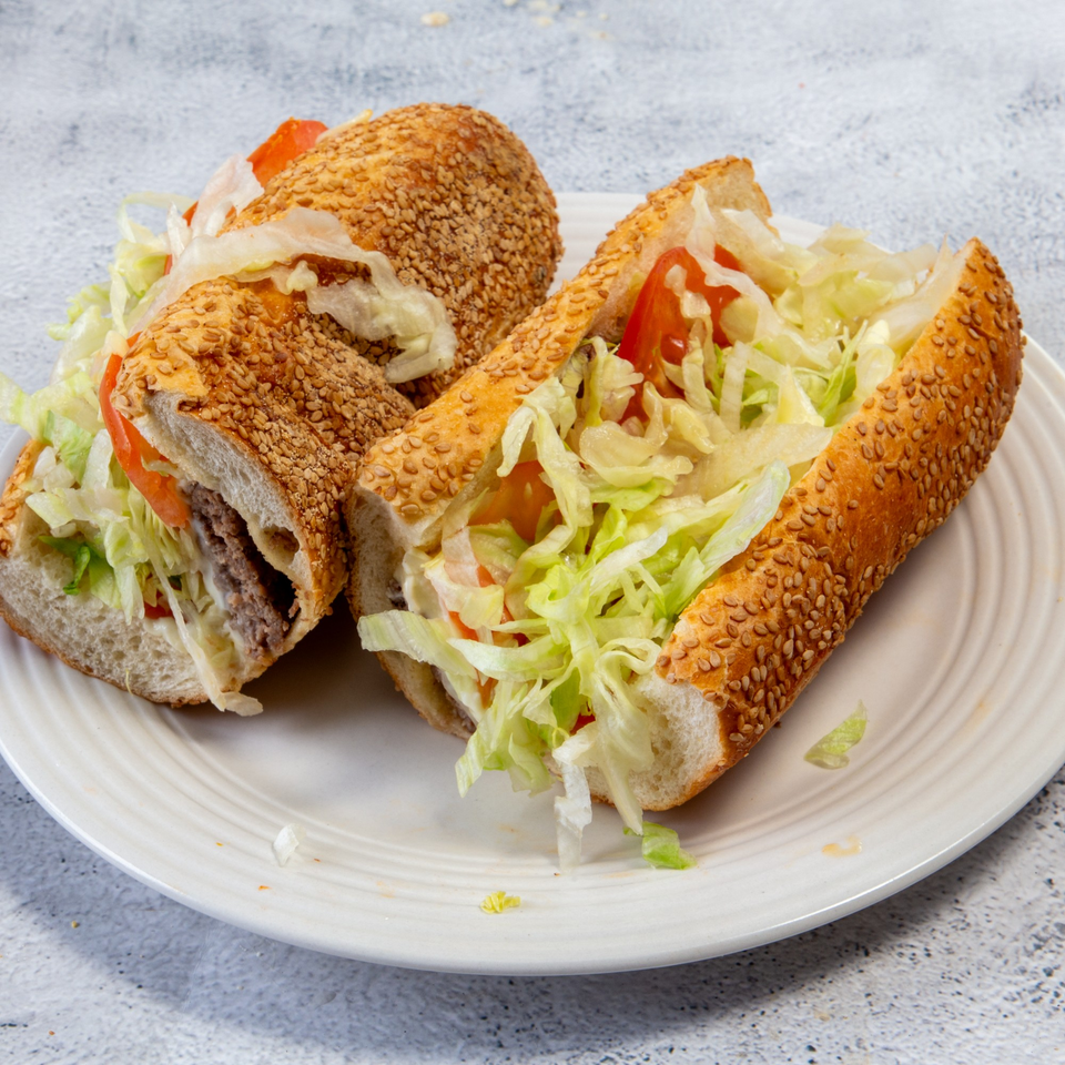 Two sesame seed hoagies filled with meat, lettuce, tomatoes, and onions on a white plate.