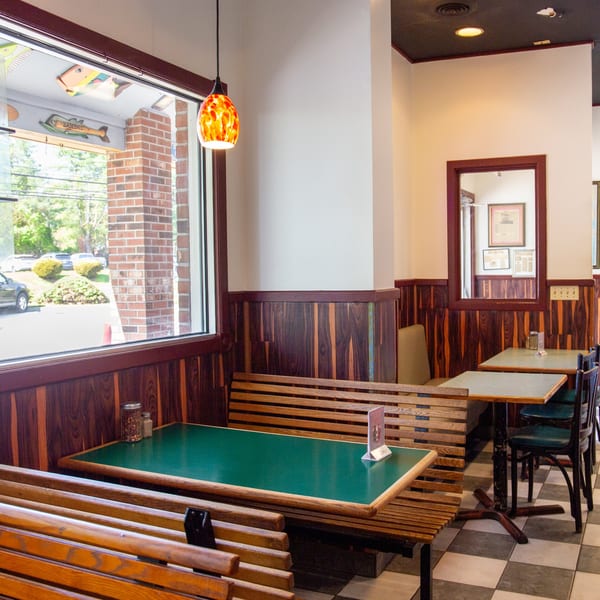 Restaurant interior with wooden booths, green-topped tables, checkered floor, and a large window overlooking a parking lot.