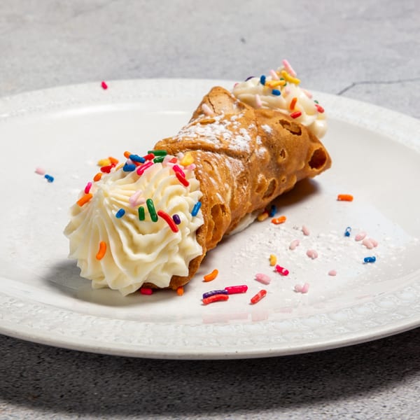 A cannoli filled with white cream and topped with colorful sprinkles and powdered sugar on a white plate.
