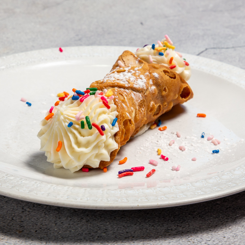 A cannoli filled with white cream and topped with colorful sprinkles and powdered sugar on a white plate.