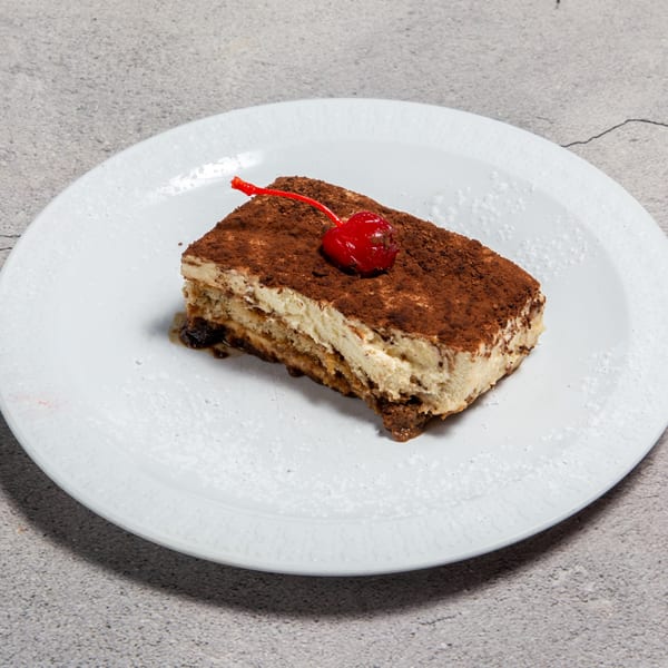 A slice of tiramisu with cocoa powder and a red cherry on a white plate.